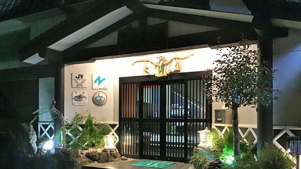 Entrada de um edifício com portão preto, plantas em vasos e sinalização iluminada.