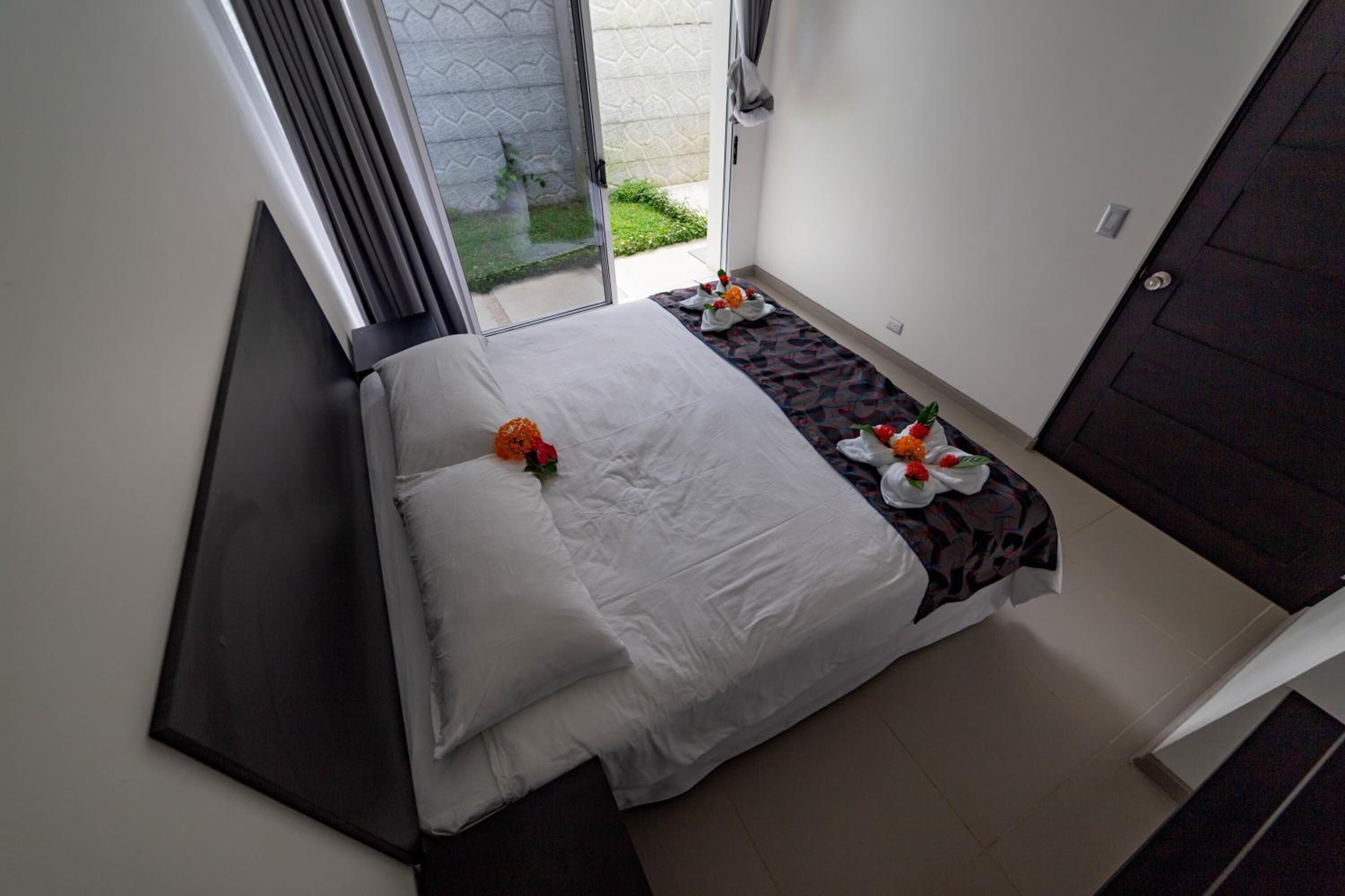 A small, neatly made bed with a white comforter and pillows, a small table with a floral arrangement, and a view of a garden through a glass door.