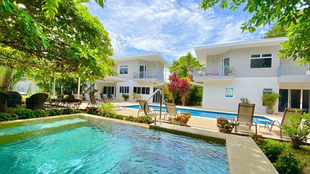 A pool area with lounge chairs and a clear blue pool, surrounded by white buildings and lush greenery. - Uvita Paradise (Bahía Ballena)