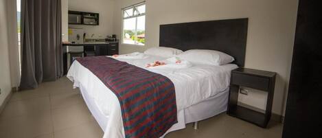 A bedroom with a bed, a bedside table, a desk with a chair, and a window.