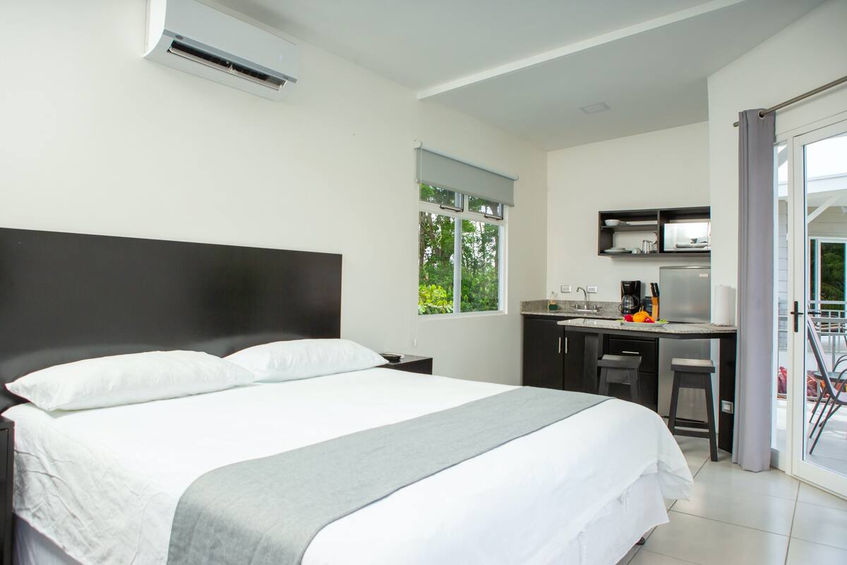 A bedroom with a large bed, a small kitchenette, and a sliding glass door to the outside.