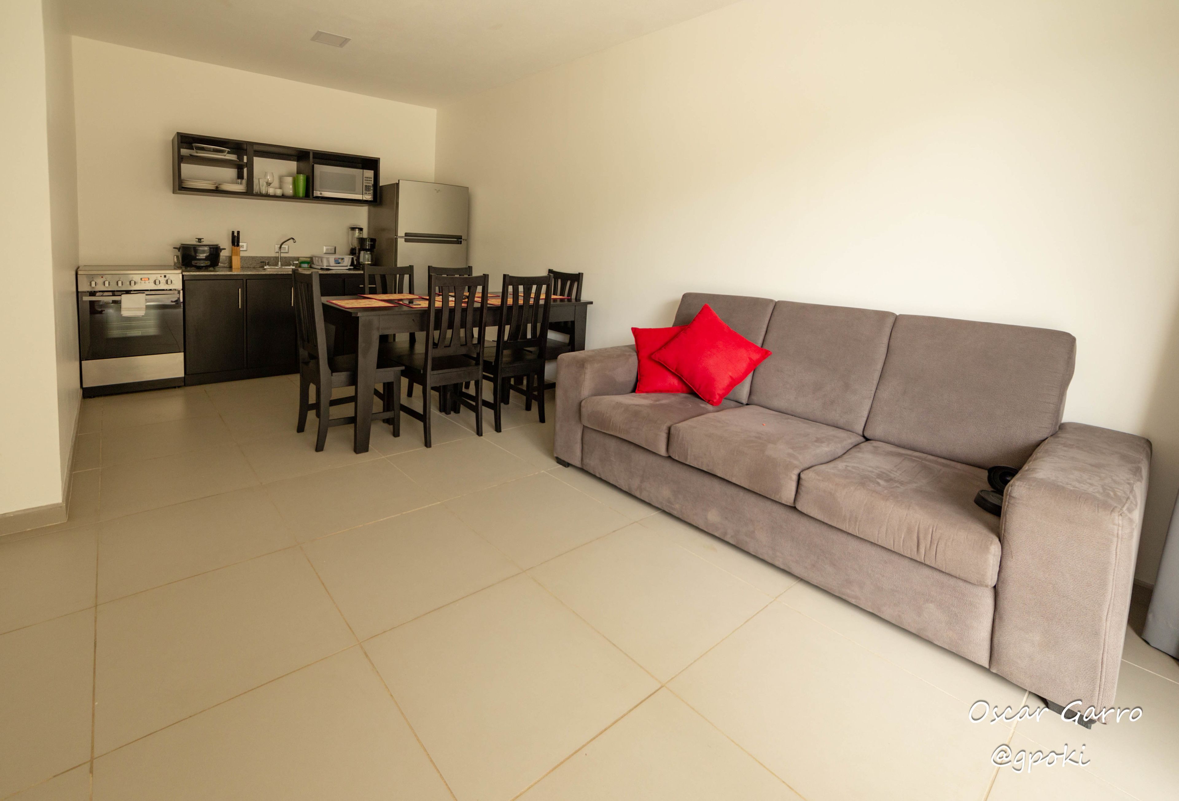 A modern kitchen with a dining area, a grey sofa with red cushions, and a tiled floor.