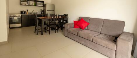 A modern kitchen with a dining area, a grey sofa with red cushions, and a tiled floor.