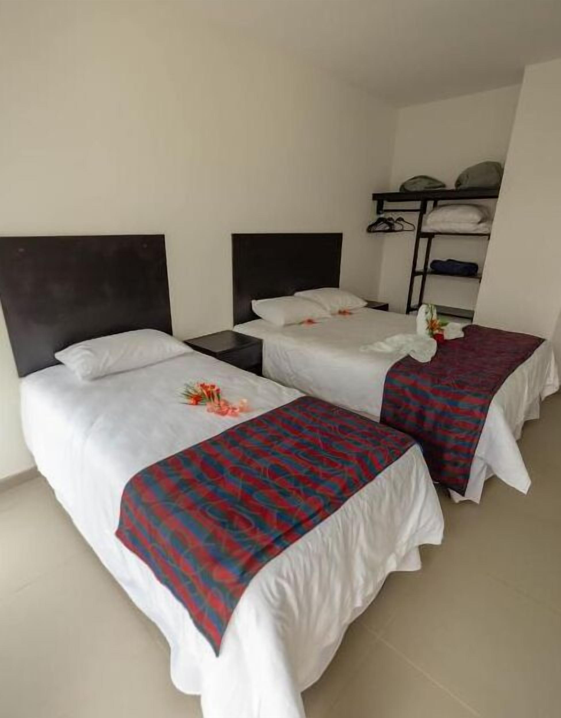 A room with two beds, each with white linens and a red patterned blanket. There is a shelf with folded clothes and a small plant on the wall.
