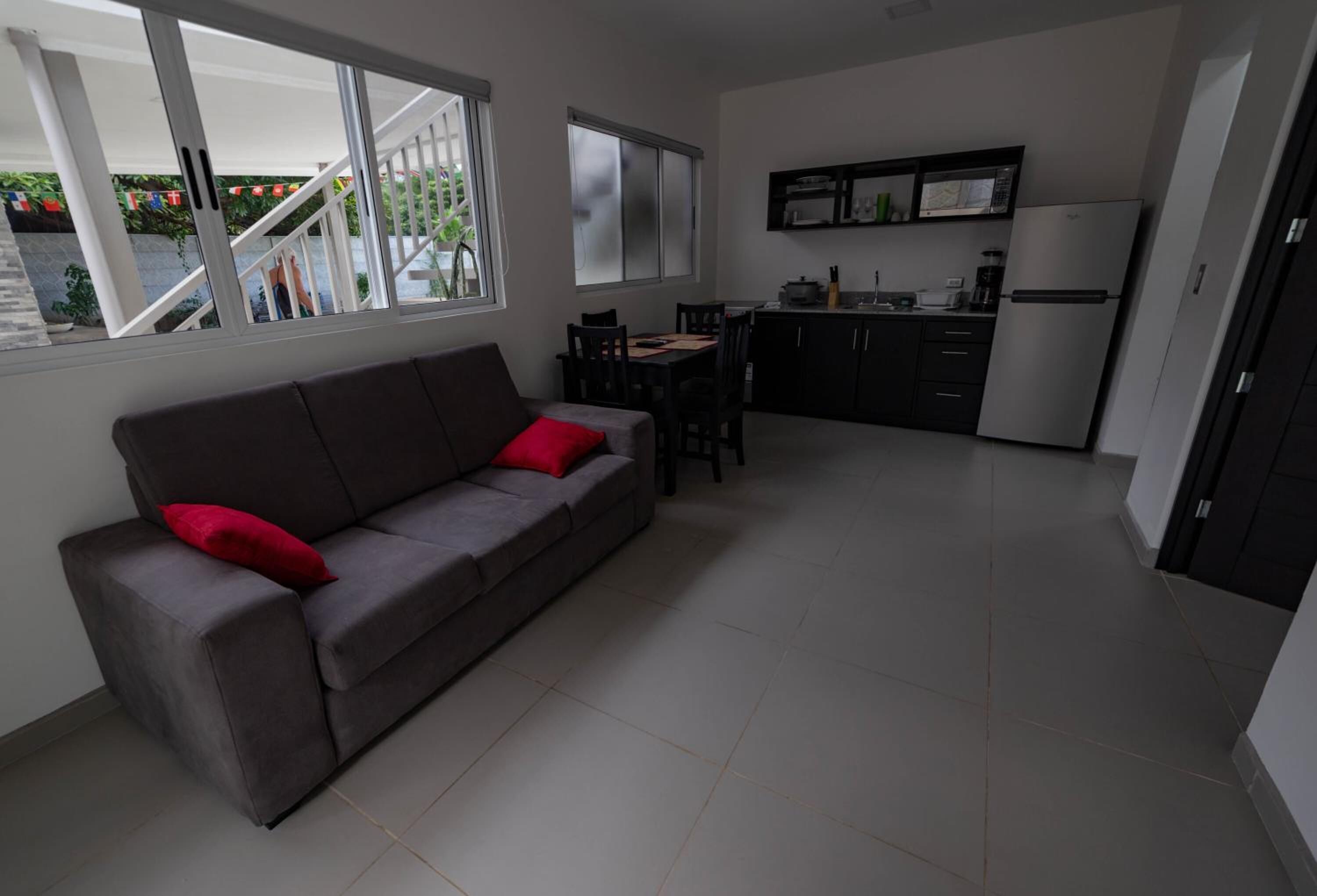 A living room with a grey sofa, red cushions, a dining area with a table and chairs, and a kitchen with cabinets and appliances.