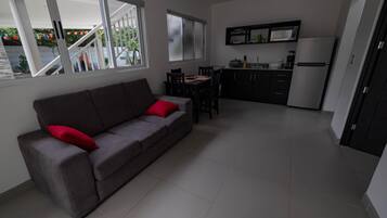 A living room with a grey sofa, red cushions, a dining area with a table and chairs, and a kitchen with cabinets and appliances.