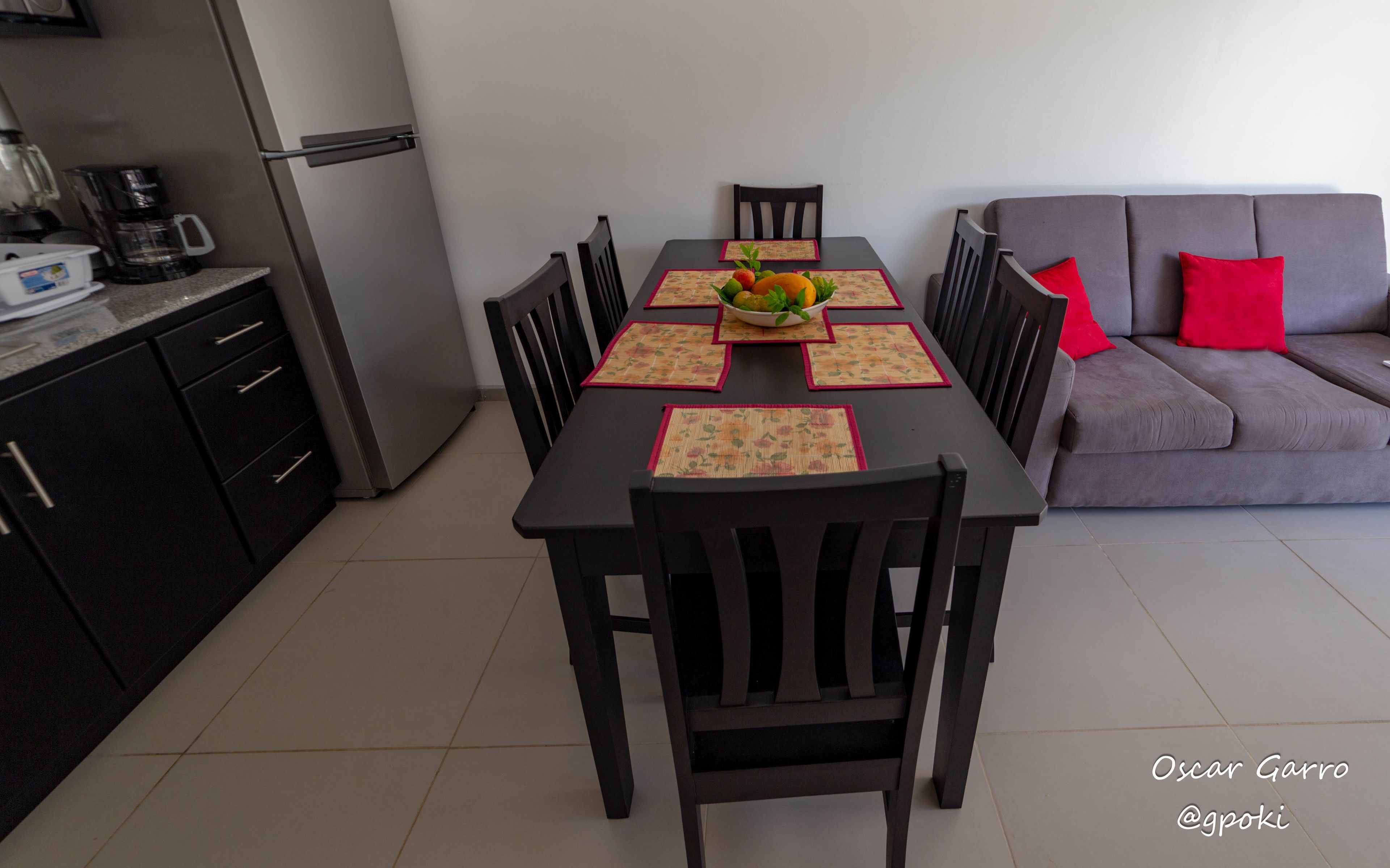 A dining area with a table set for four, surrounded by chairs, and a grey sofa with red cushions in the background.