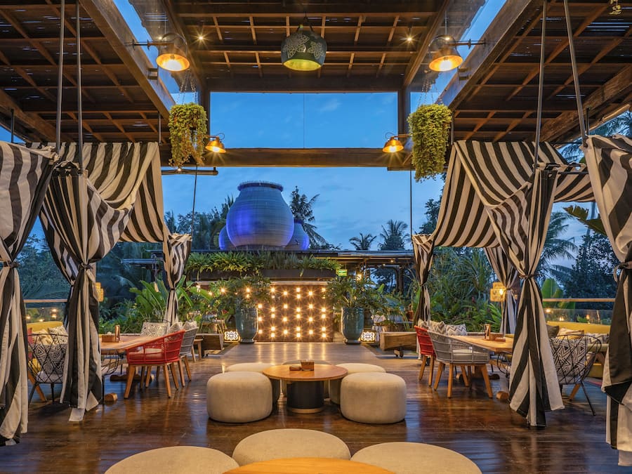 An outdoor dining area with round tables and chairs, a wooden ceiling, and hanging plants.