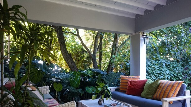 A covered outdoor seating area with a wicker sofa, a wooden coffee table, and various plants. - Africa's Eden Guesthouse (Pietermaritzburg)