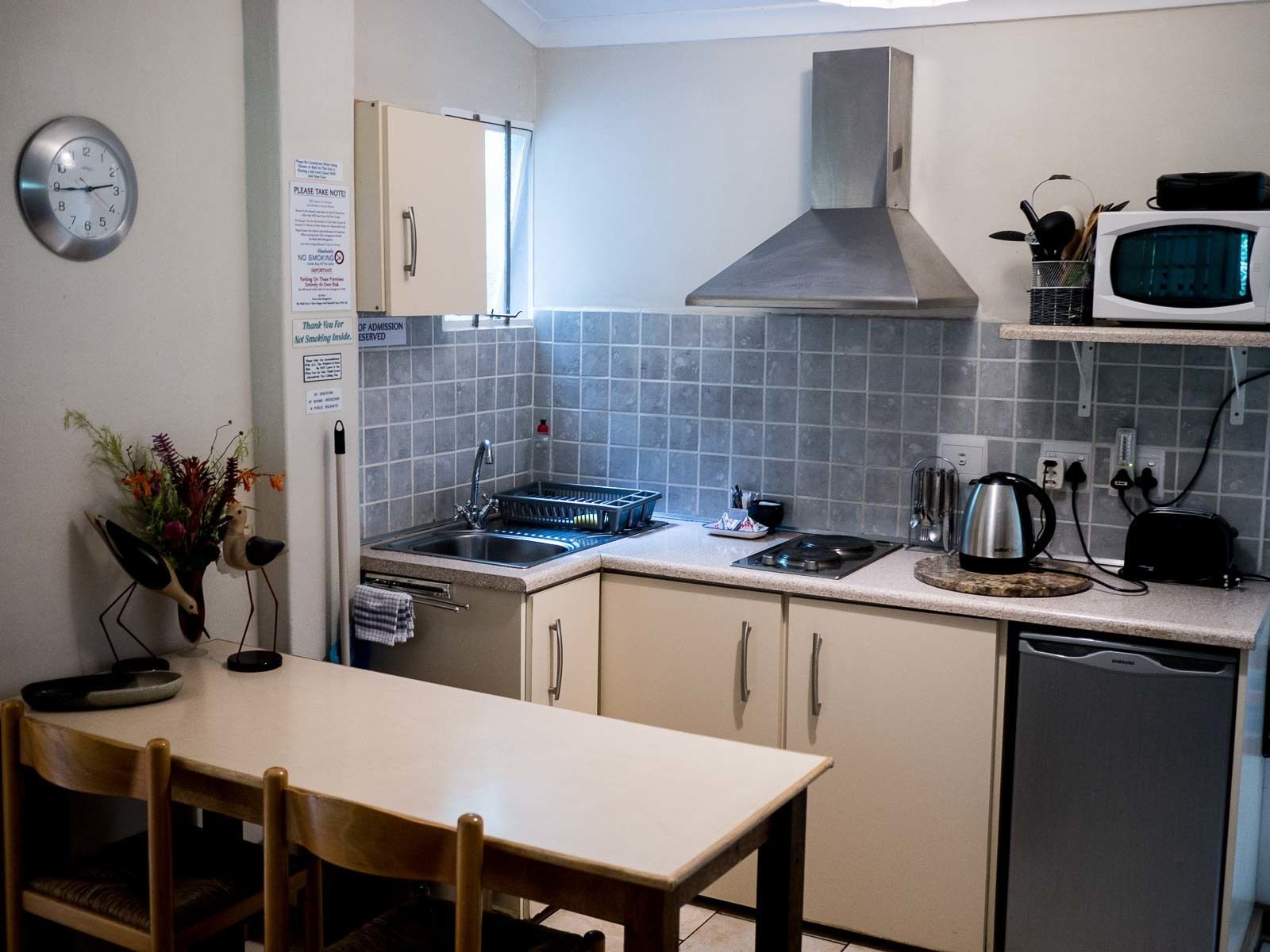 A compact kitchen with a microwave, stove, sink, and dishwasher. There is a clock on the wall and a sign indicating no smoking.