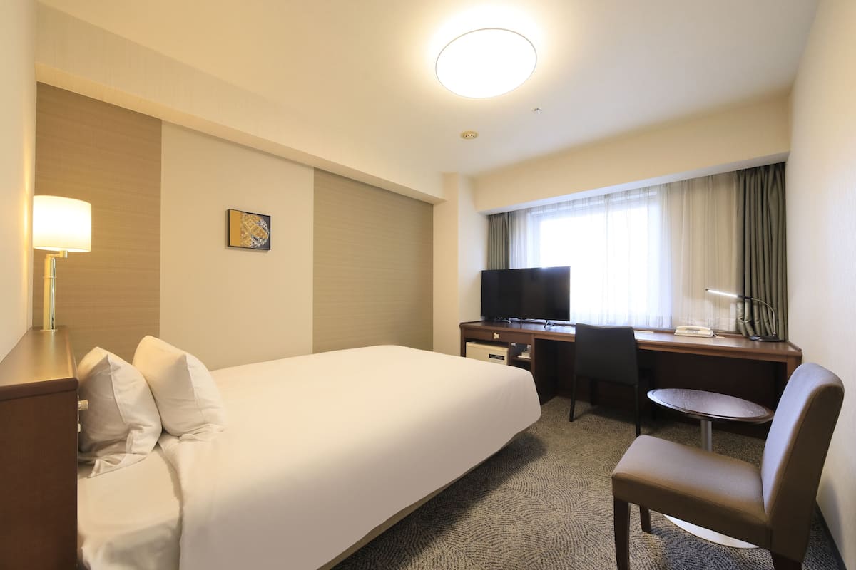 A hotel room with a bed, desk, chair, TV, and a lamp.
