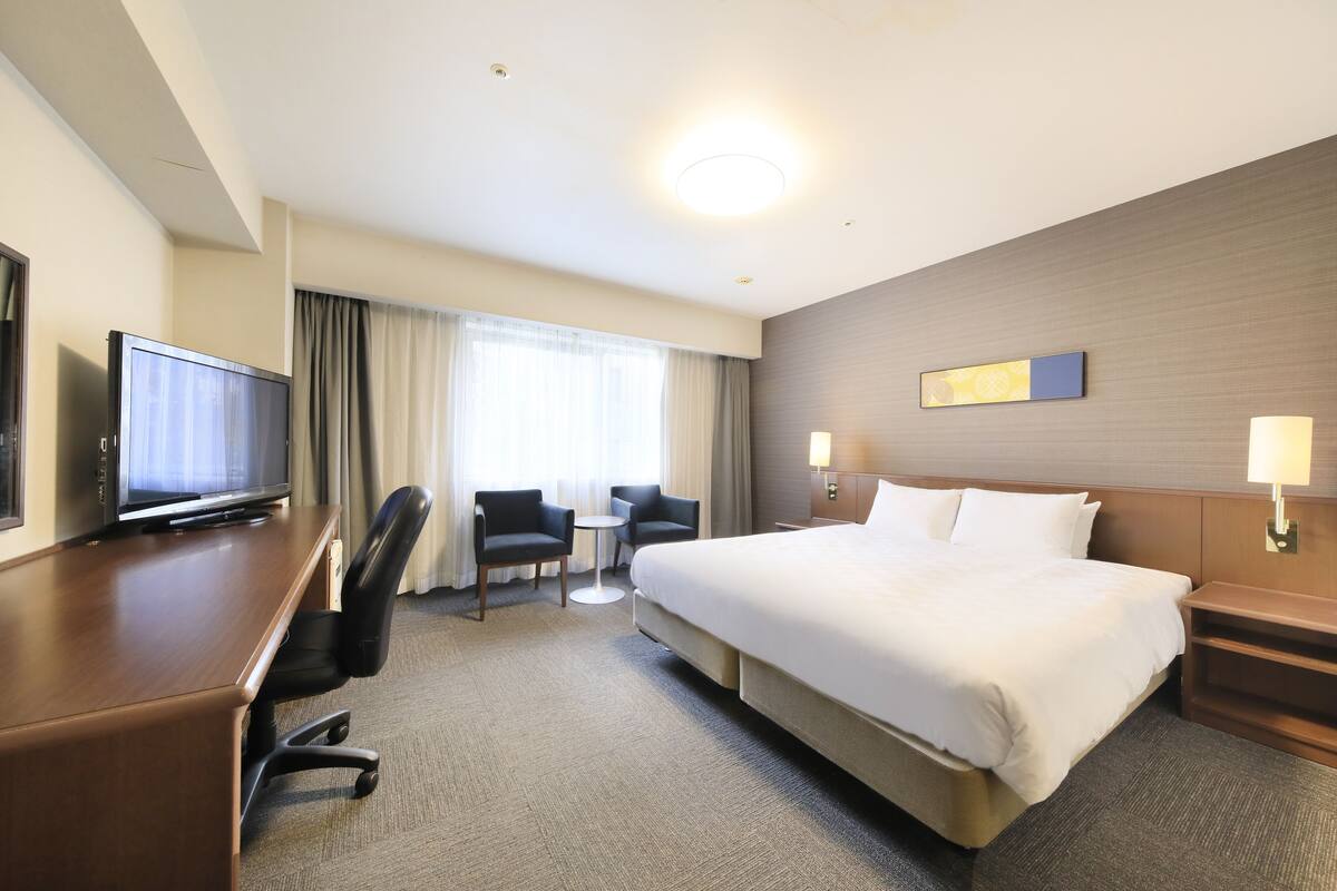A hotel room with a large bed, a desk with a television, two armchairs, a small table, and a window with curtains.