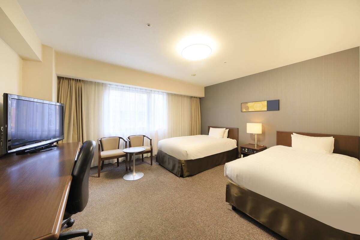 A hotel room with a large bed, a smaller bed, a desk with a television, a chair, and a lamp.