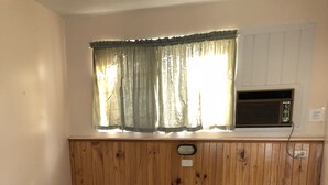 Un lit simple avec une tête de lit en bois, deux tables de chevet, une fenêtre avec des rideaux et un appareil de climatisation intégré.