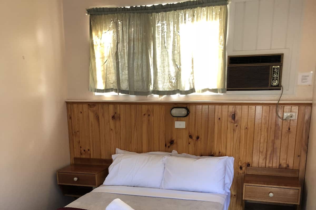 A single bed with a wooden headboard, two bedside tables, a window with curtains, and a built-in air conditioning unit.
