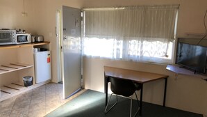 Une pièce de taille modeste comprenant un coin cuisine, une chaise, une petite table, un téléviseur et une fenêtre ornée de rideaux légers.