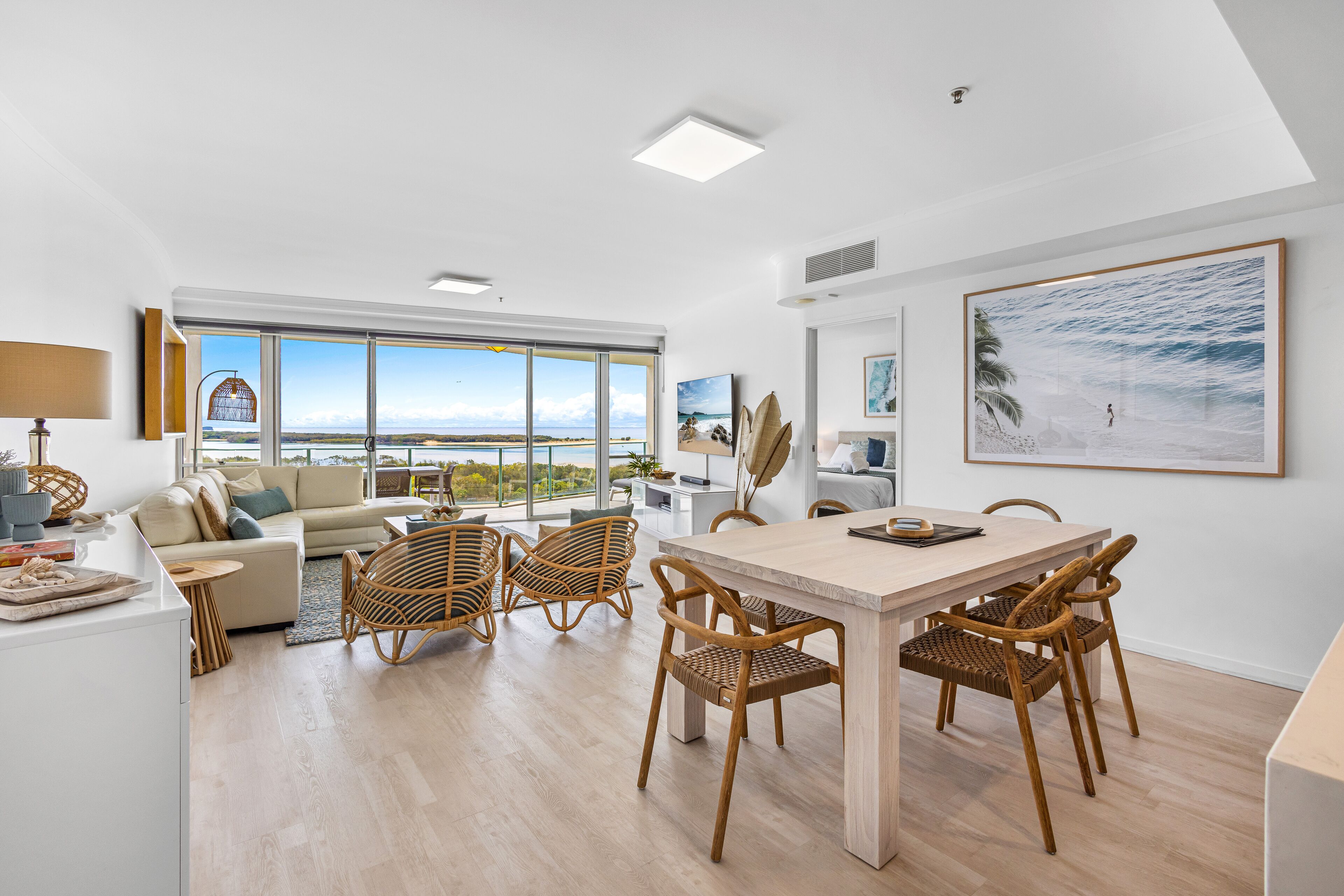 A modern living room with a large window, a dining area with wicker chairs, and a view of the ocean.