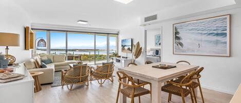 A modern living room with a large window, a dining area with wicker chairs, and a view of the ocean.