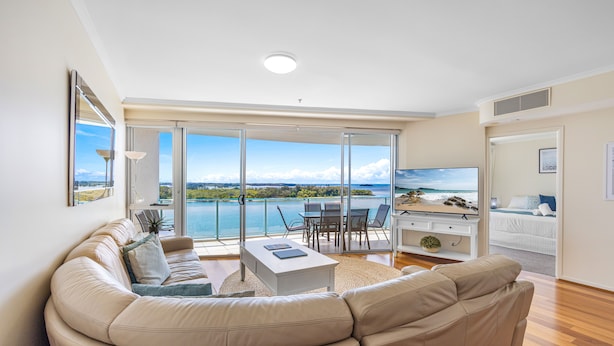 A modern living room with a large sofa, a coffee table, and a TV. There is a balcony with a view of the sea. - The Duporth Riverside (Maroochydore)