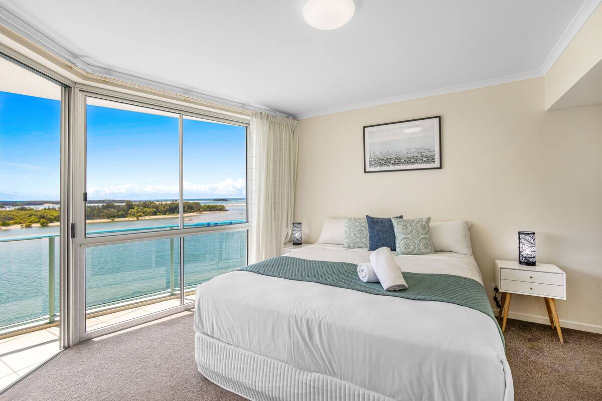 A hotel room with a large bed, a nightstand, a framed picture, and a view of the sea through a sliding glass door.