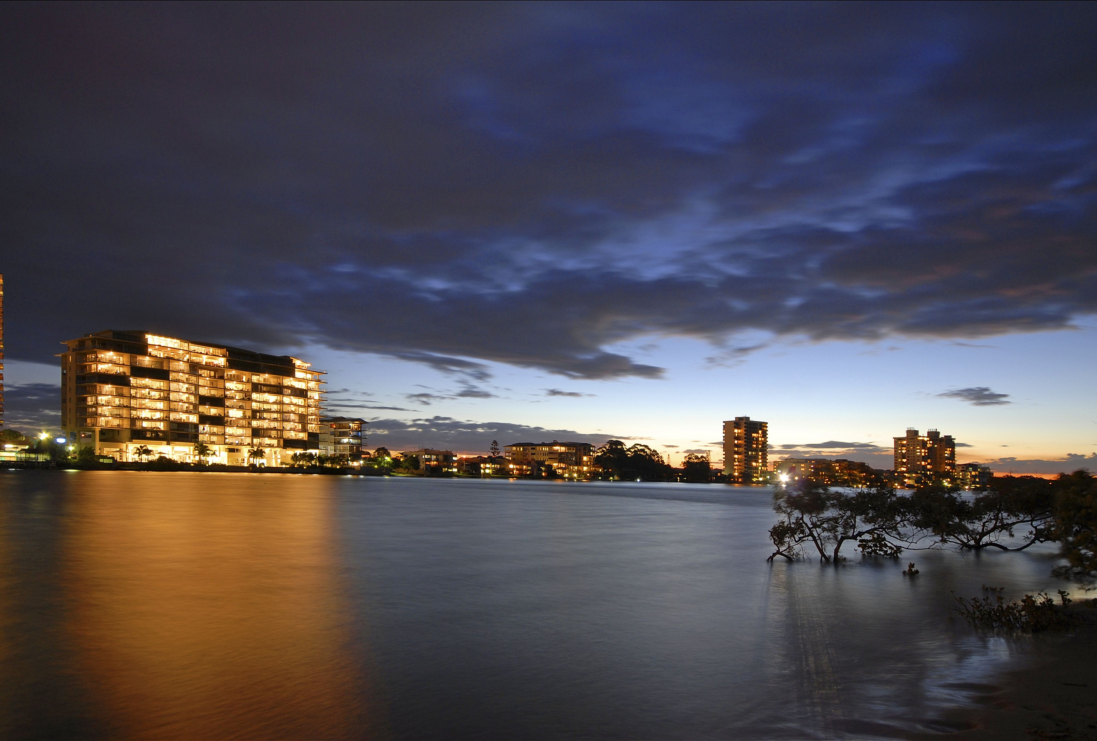 Un hotel frente al mar con varios pisos y balcones, un cuerpo de agua tranquilo en primer plano y un horizonte urbano al fondo.