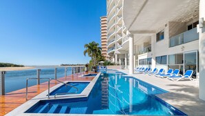A swimming pool with a view of the beach, lounge chairs, and a tall building with balconies. - The Duporth Riverside (Maroochydore)