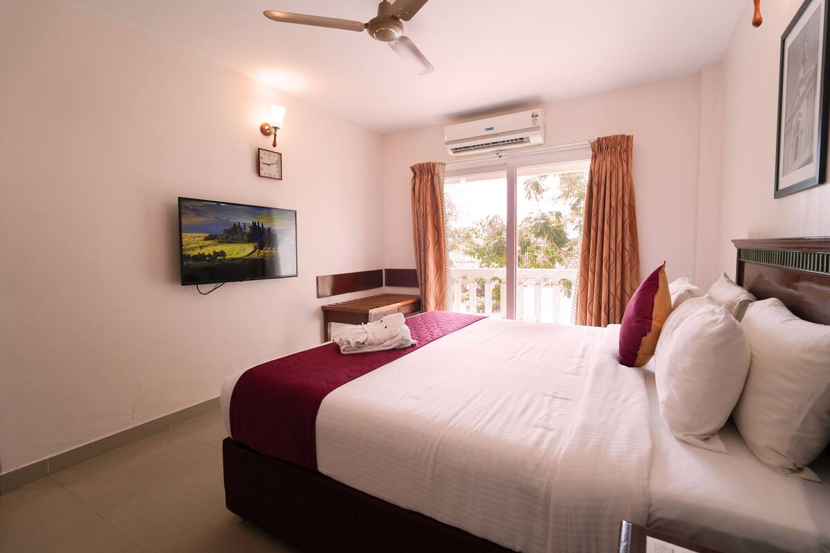 Ein Hotelzimmer mit Bett, Fernseher, Deckenventilator, Klimaanlage, einem Fenster mit Vorhängen und einer Wanduhr.