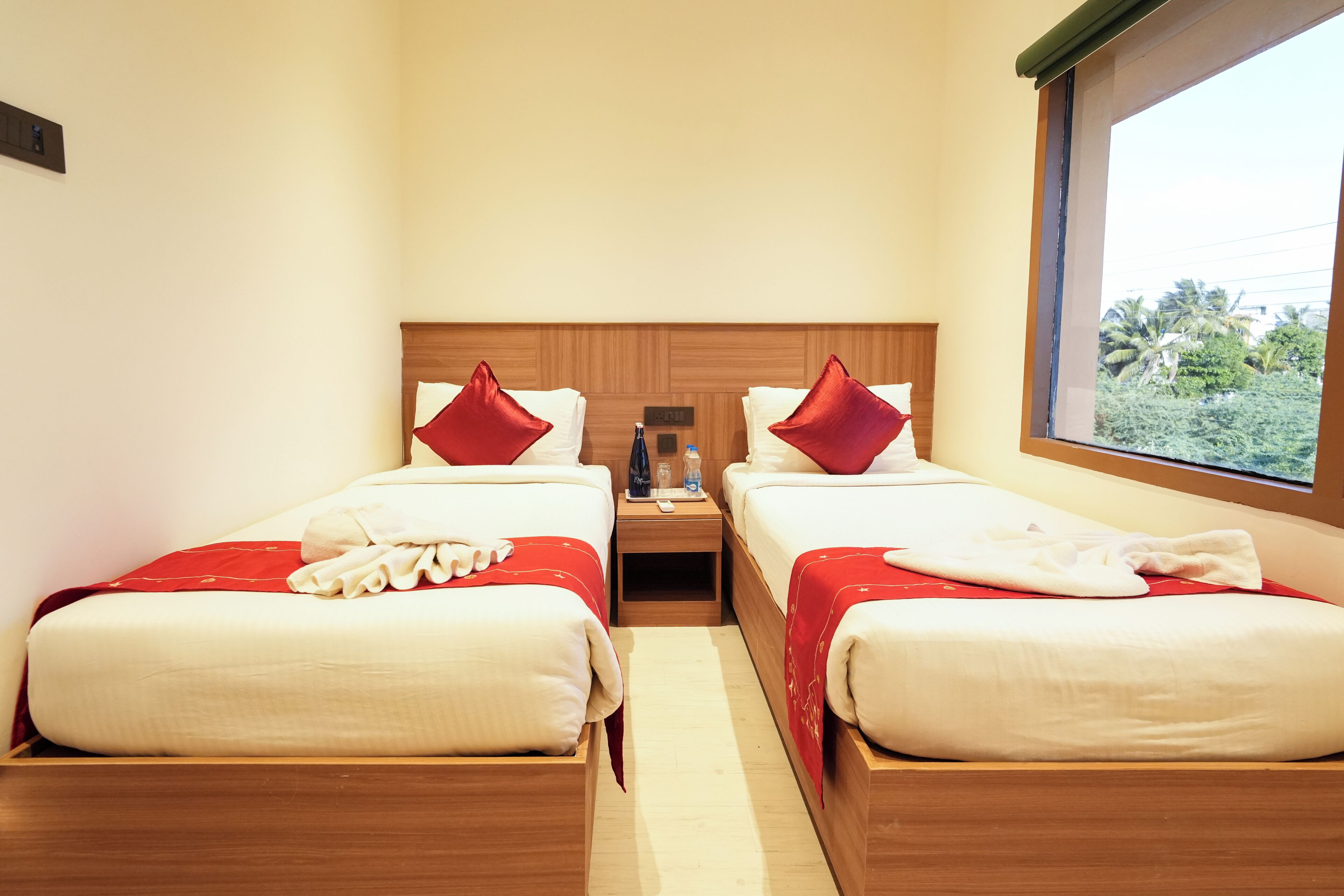 A hotel room with two beds, each with a red pillow and white bedding. There is a wooden headboard, a nightstand with a bottle and a remote control, and a window with a view of greenery outside.