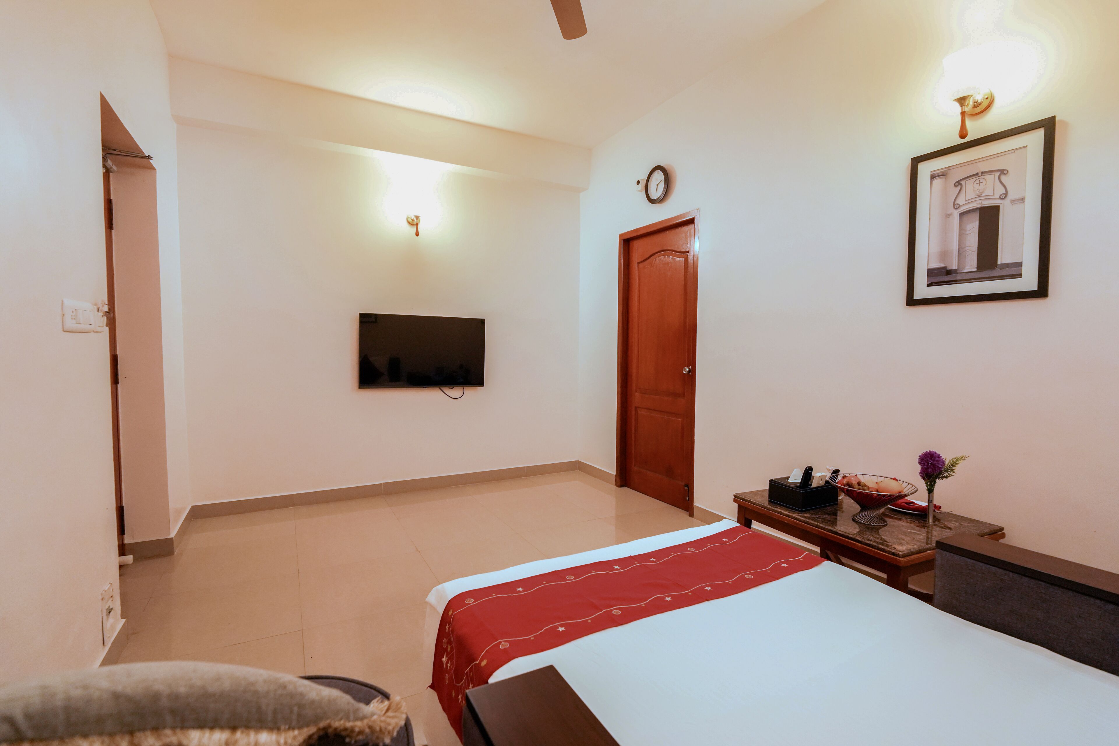 A hotel room with a bed, a TV mounted on the wall, a wooden door, and a small table with a bowl and a flower.