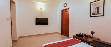 A hotel room with a bed, a TV mounted on the wall, a wooden door, and a small table with a bowl and a flower.