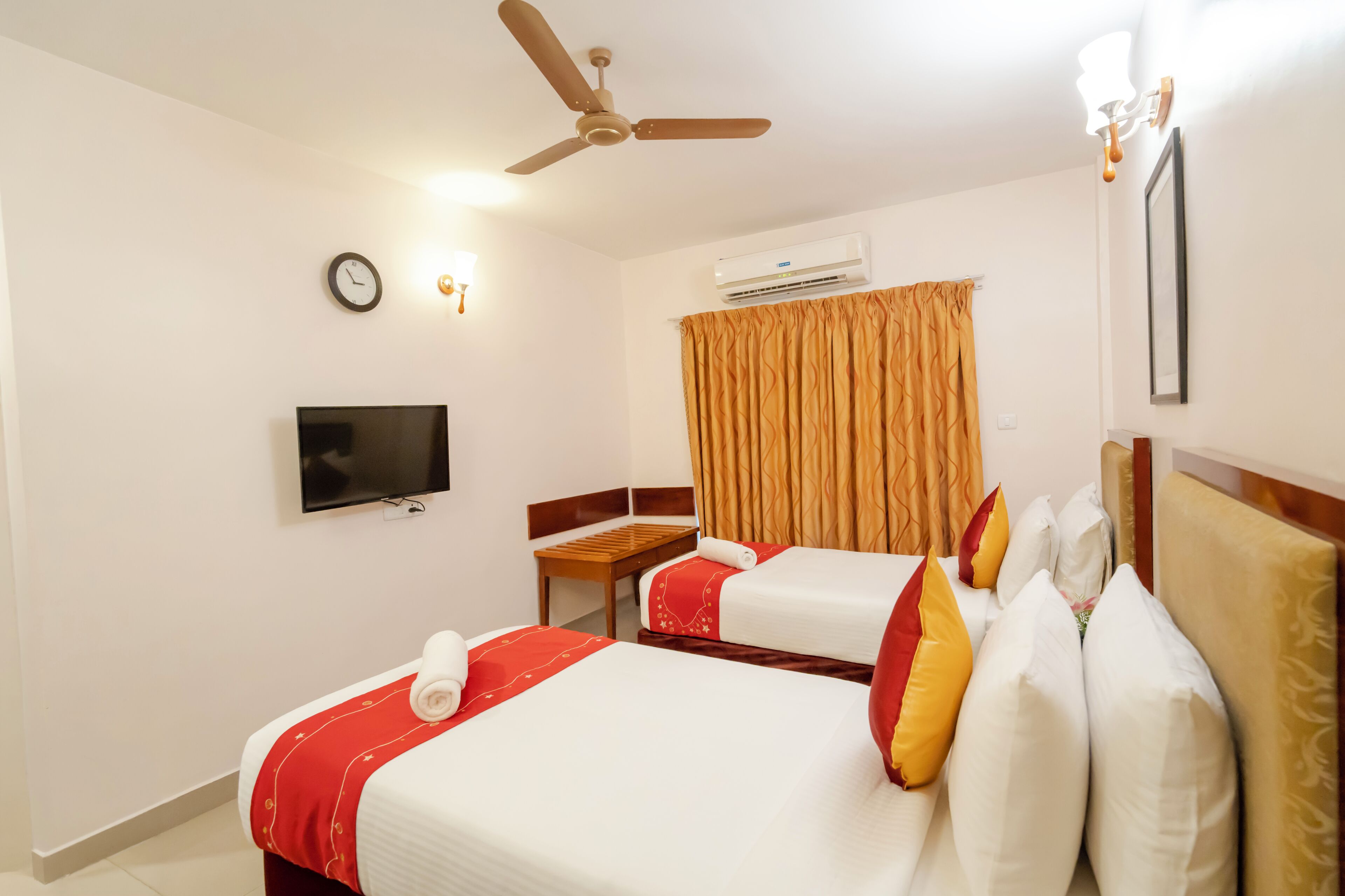 A hotel room with two beds, a television, a ceiling fan, an air conditioning unit, a clock, and a window with curtains.