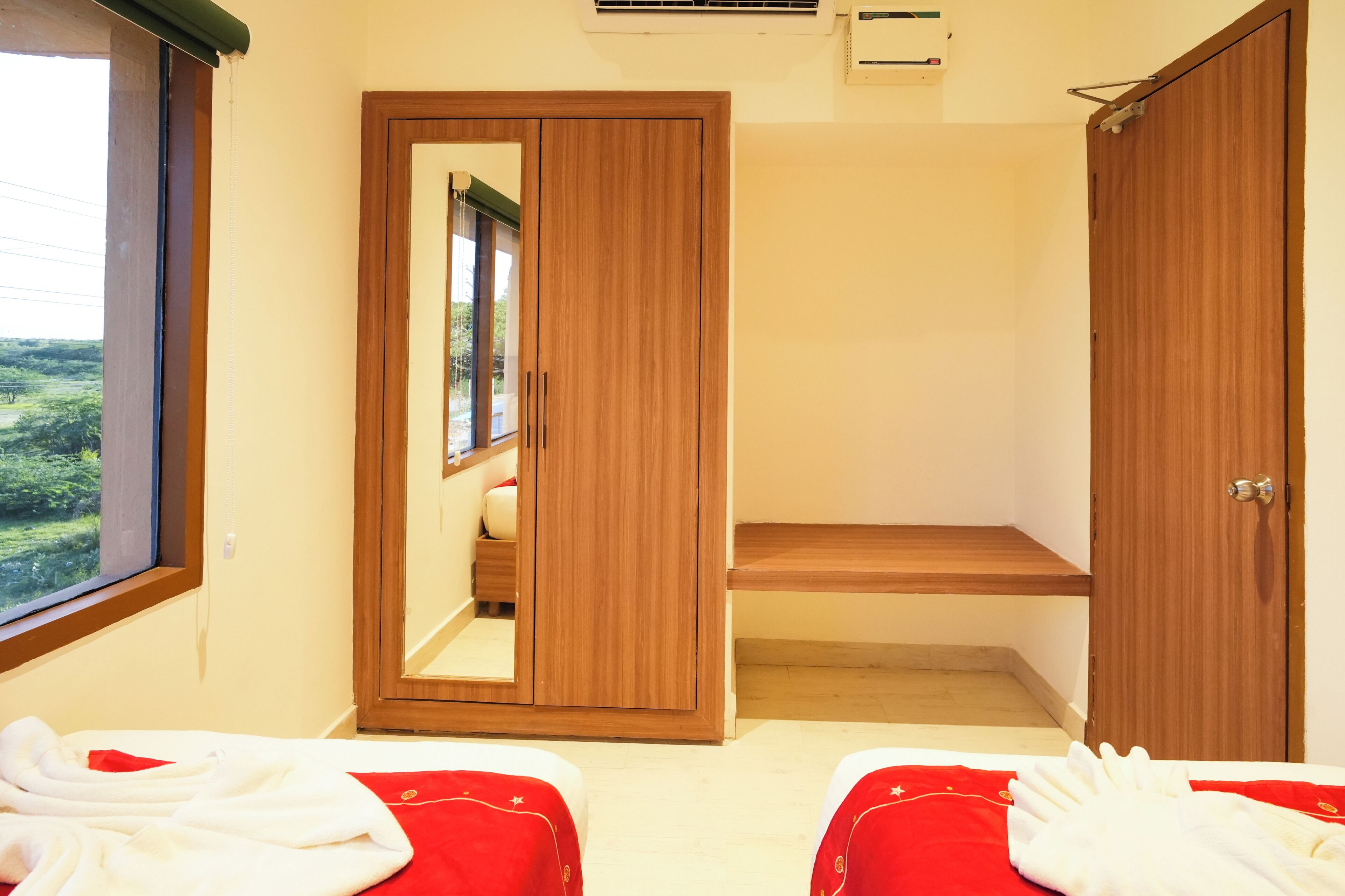 A room with a wooden wardrobe, a mirror, a window with a view, and two beds with red headboards.