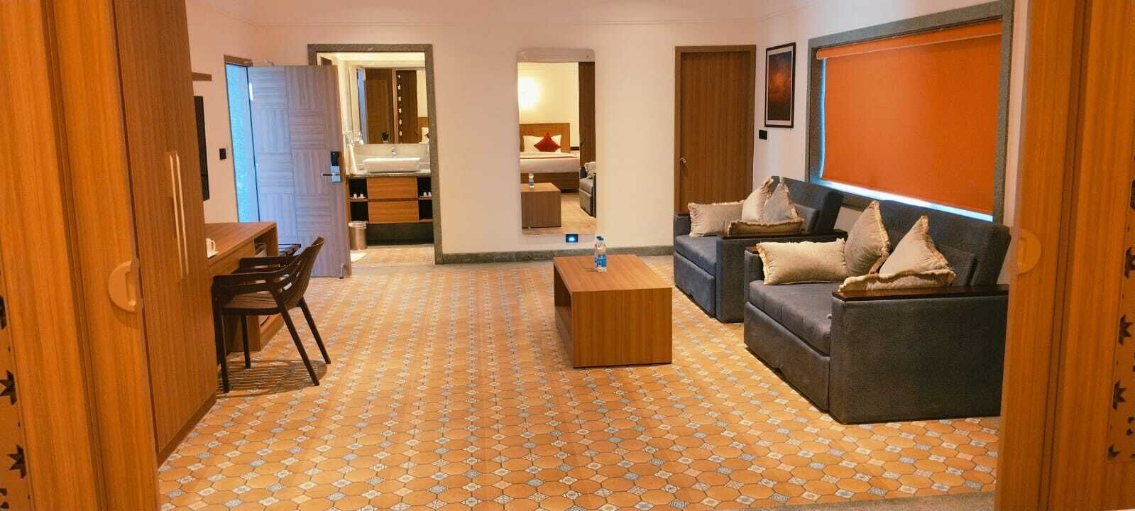 Habitación de hotel con sofá, mesa de centro, televisor, espejo y baño visible a través de una puerta abierta.