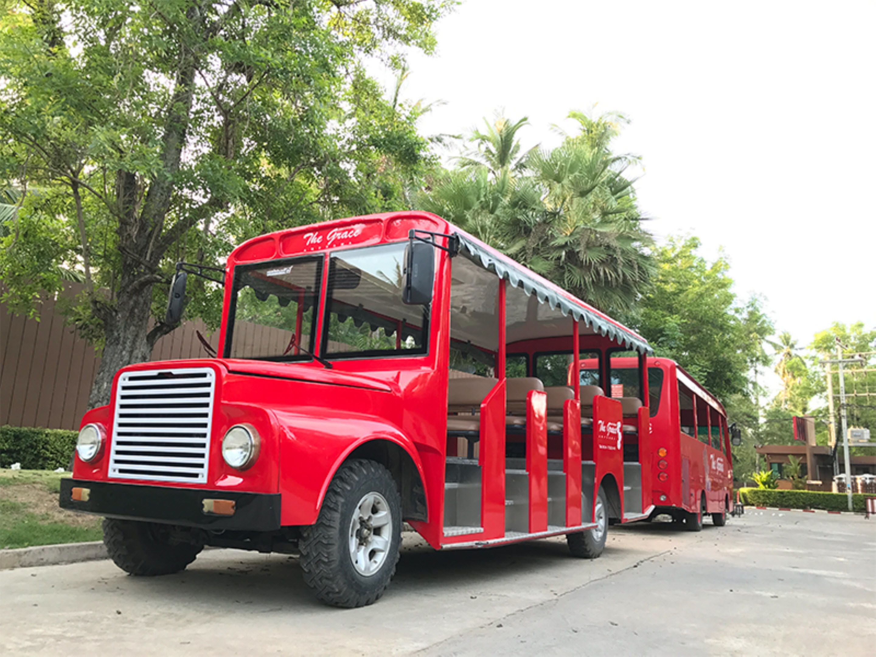 Um autocarro vermelho antigo com a frente coberta e os lados abertos, estacionado numa estrada pavimentada com árvores e um edifício ao fundo.