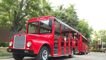 Um autocarro vermelho antigo com a frente coberta e os lados abertos, estacionado numa estrada pavimentada com árvores e um edifício ao fundo.