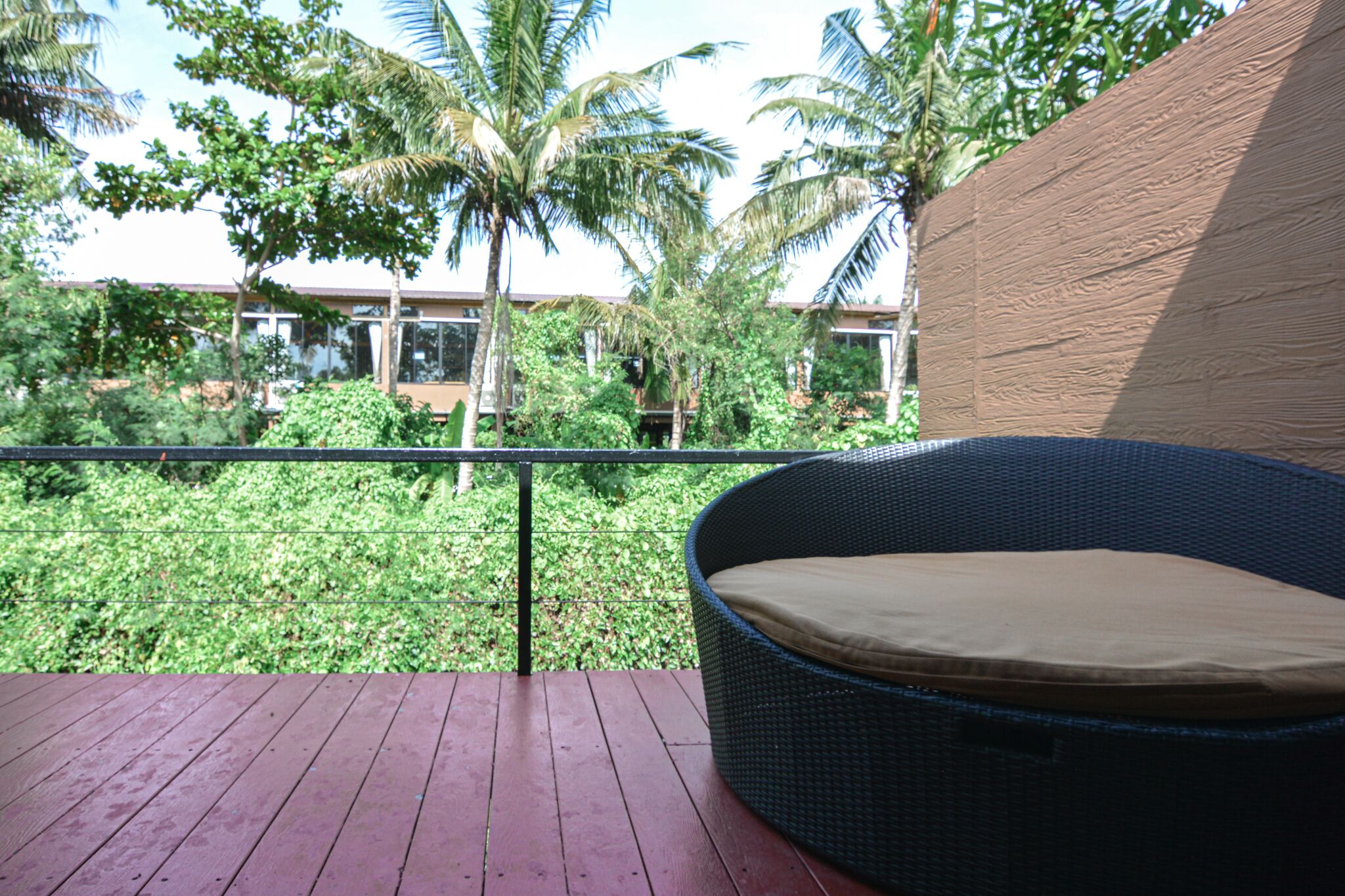 Une chaise en osier avec un coussin, placée sur une terrasse en bois, d’où l’on peut admirer un jardin luxuriant avec des palmiers et un bâtiment en arrière-plan.