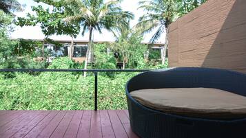 Une chaise en osier avec un coussin, placée sur une terrasse en bois, d’où l’on peut admirer un jardin luxuriant avec des palmiers et un bâtiment en arrière-plan.
