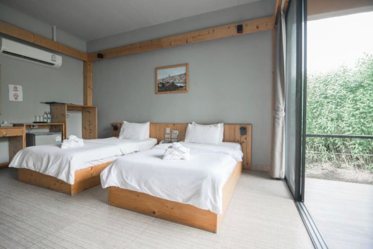 A hotel room with two beds, a wooden headboard, a small desk with a chair, and a window with a view of greenery.