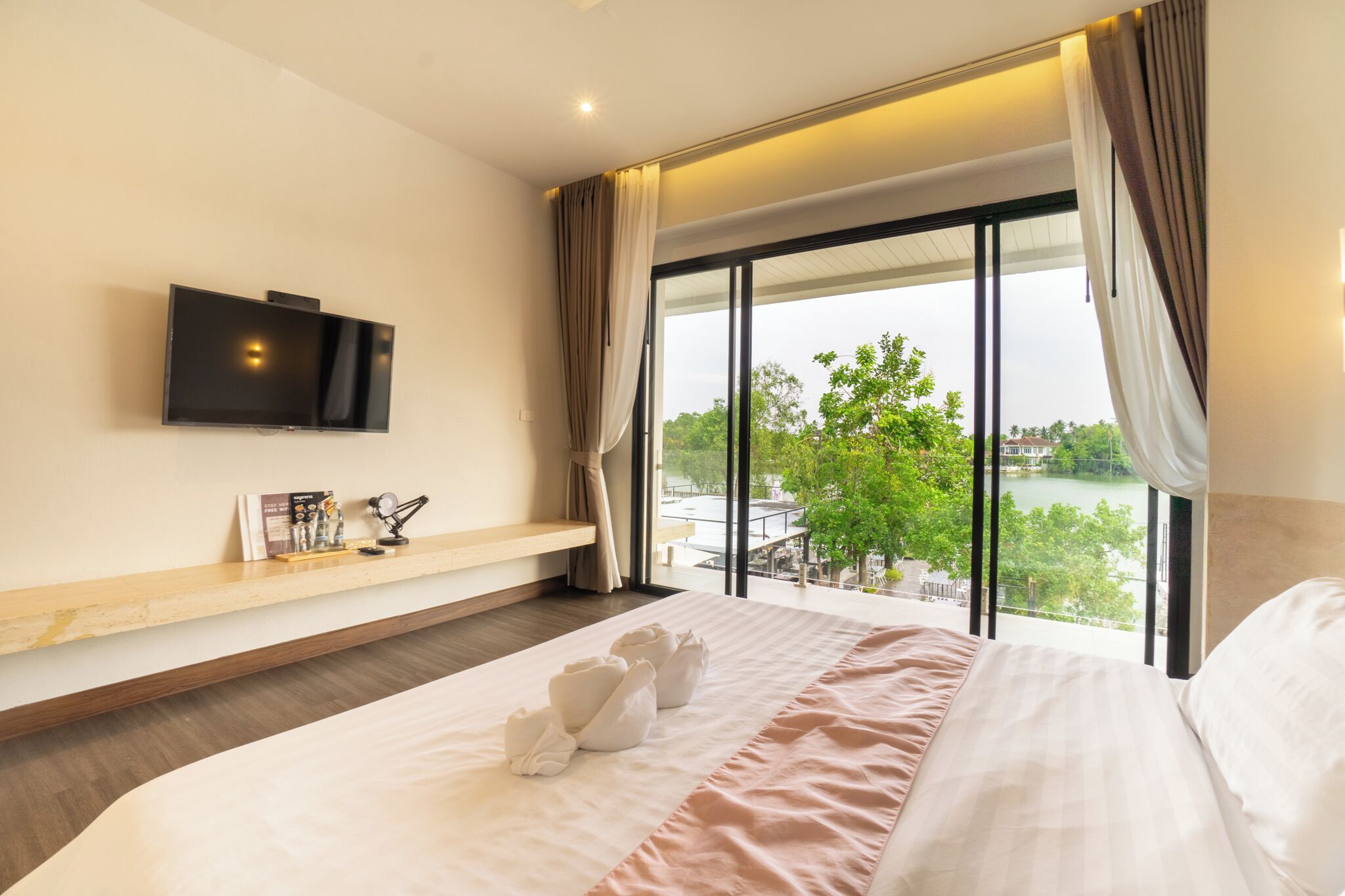 A hotel room with a bed, a TV mounted on the wall, a window with curtains, and a view of greenery outside.