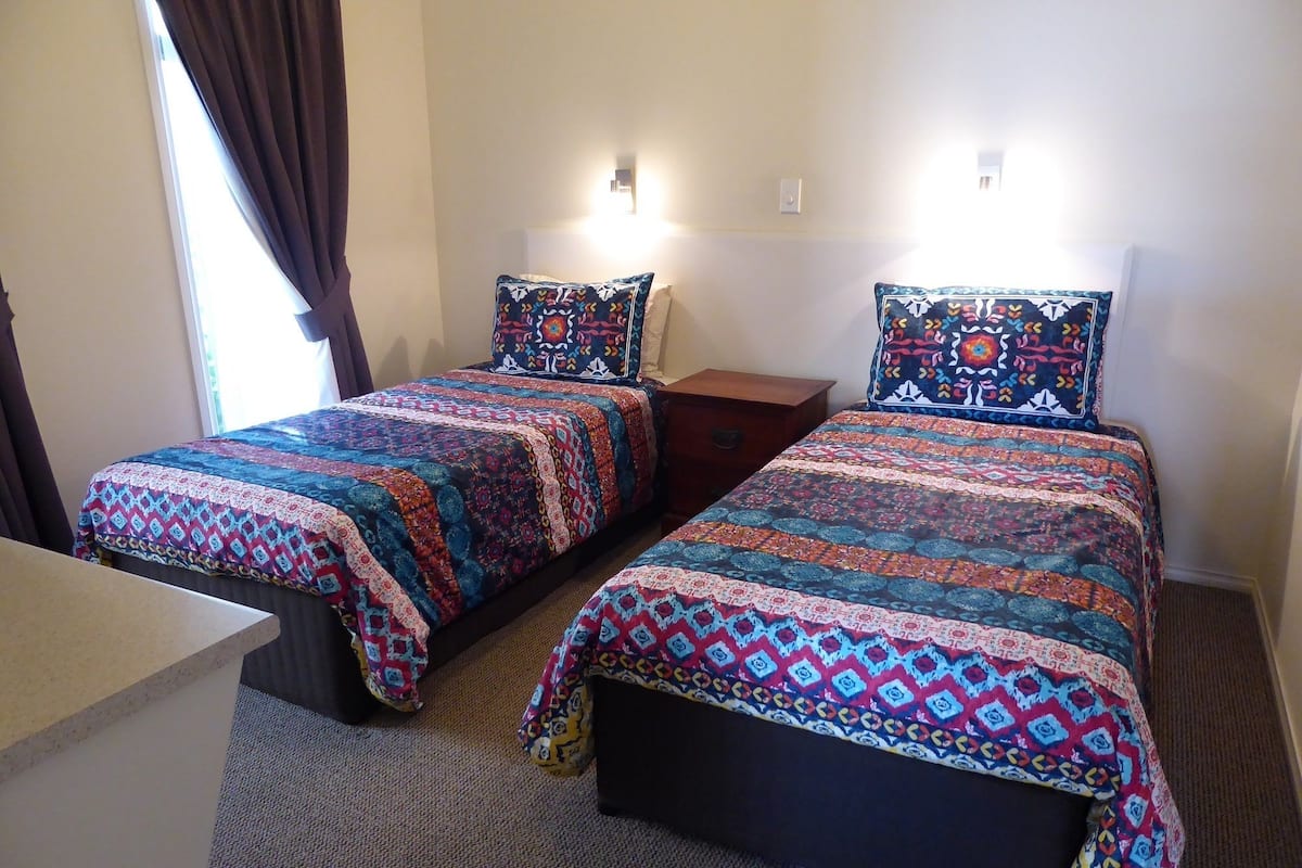 A room with two beds, each with patterned bedding and pillows, a small wooden nightstand, and a desk.
