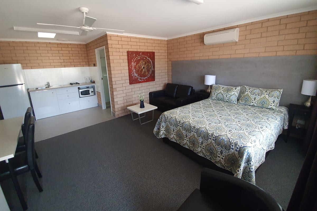 A compact hotel room with a bed, a small desk, a chair, a sofa, a kitchenette, and a ceiling fan.