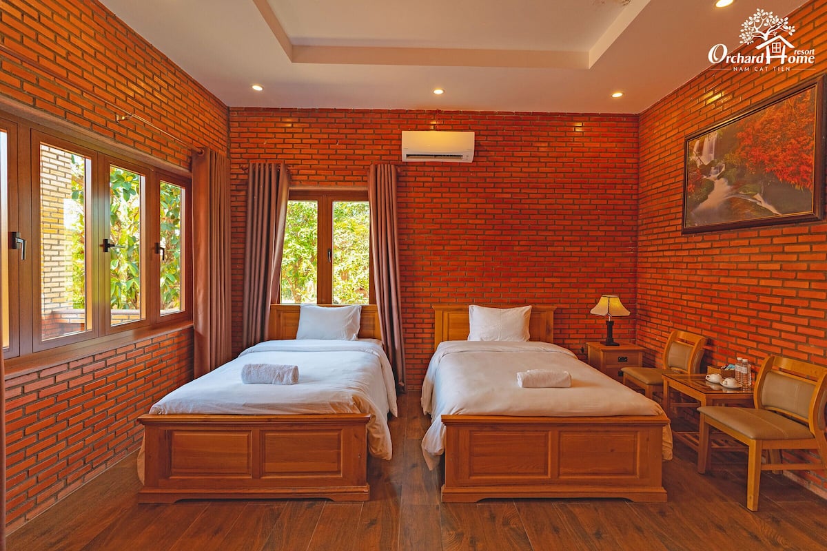 Une chambre d’hôtel avec deux lits en bois, un mur en briques, une fenêtre avec des rideaux, un climatiseur et un tableau accroché au mur.