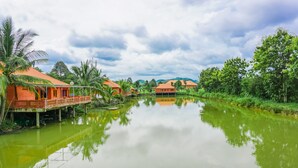 A serene riverside view with traditional-style buildings, a wooden bridge, and lush greenery.