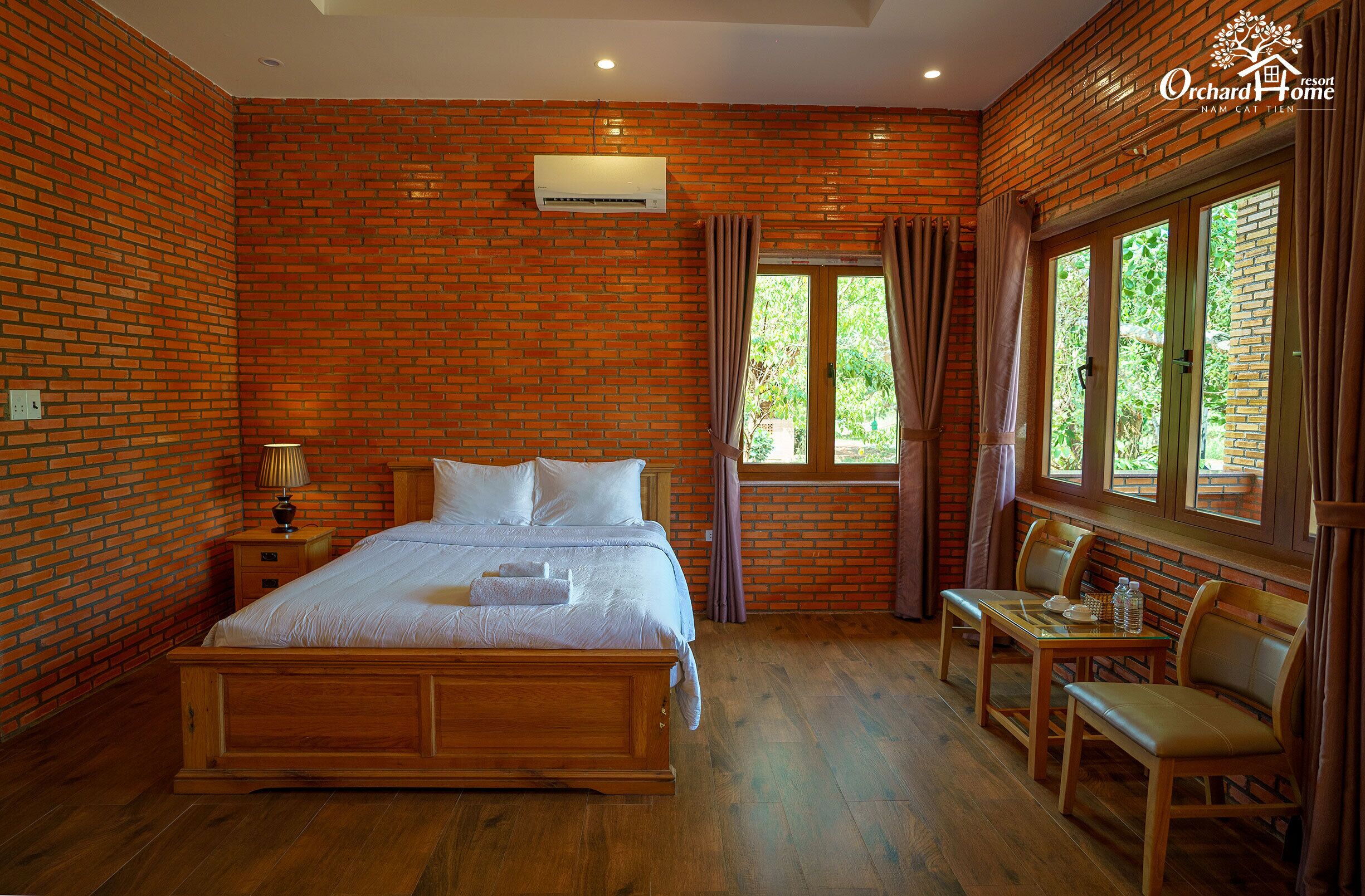 A bedroom with a brick wall, a wooden bed, a nightstand, a table with chairs, and a window with curtains.