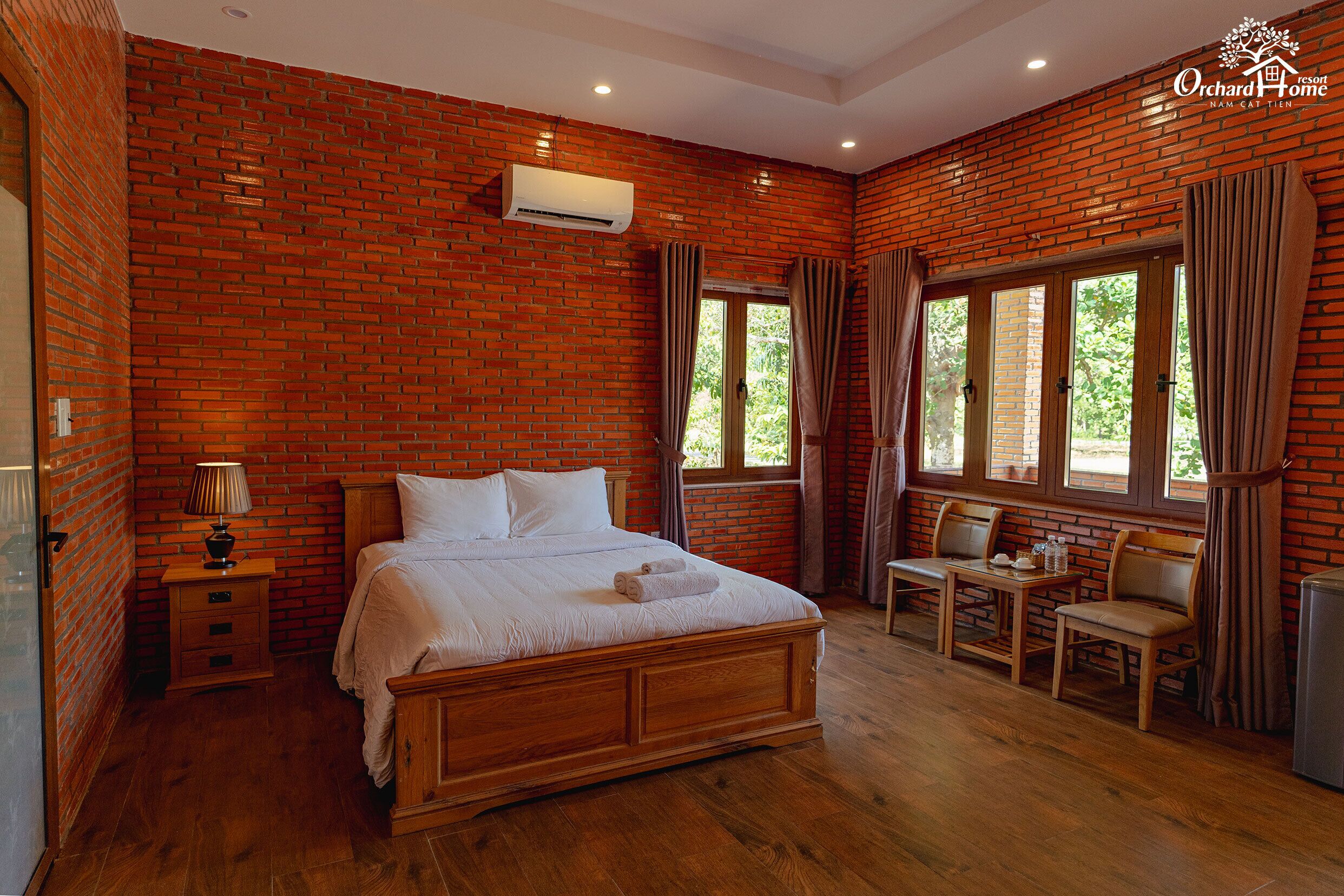 A room with a brick wall, a wooden bed, a nightstand, a table with chairs, and large windows with curtains.