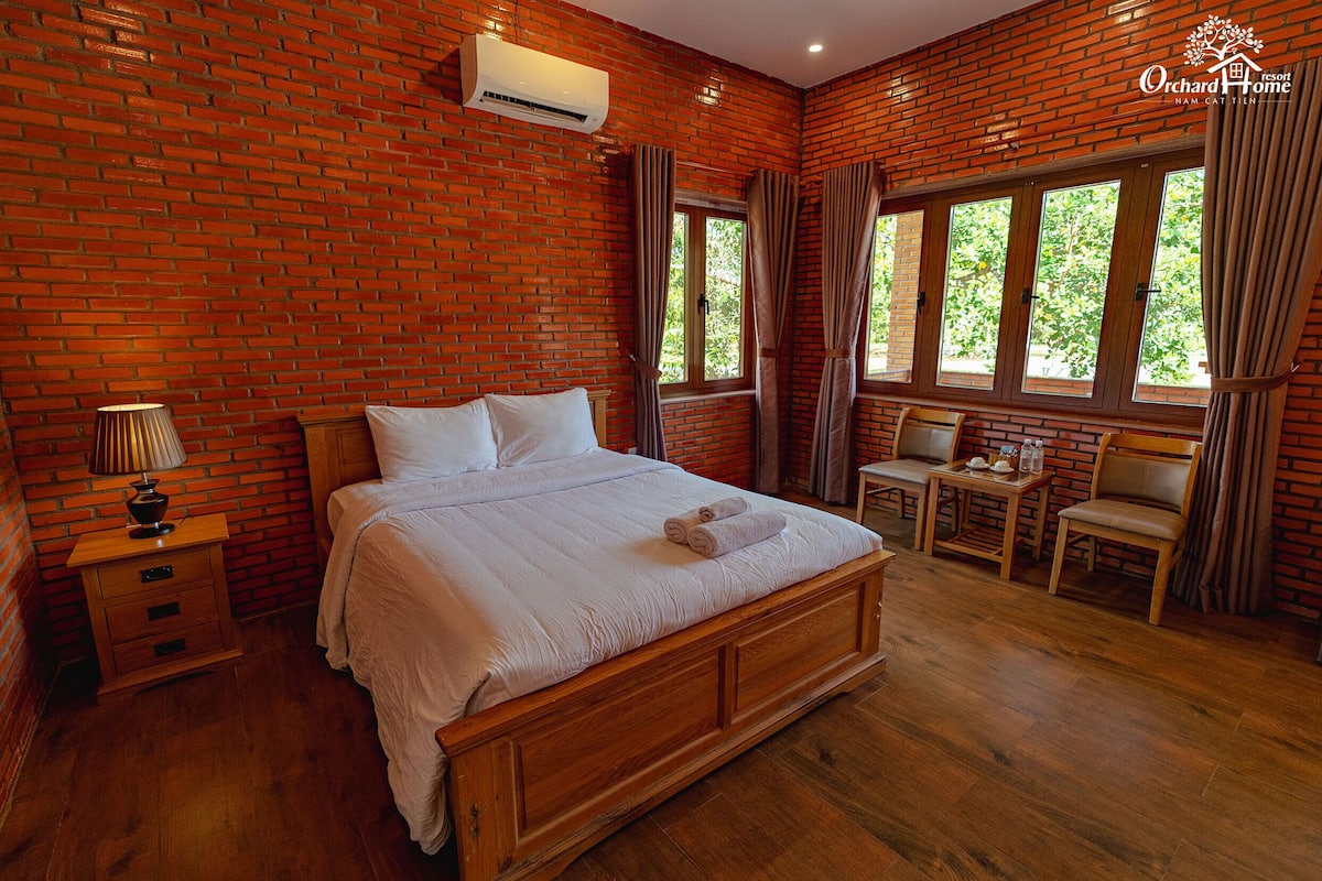 Une chambre avec un mur en briques, un lit en bois, une table de chevet, une table avec des chaises et de grandes fenêtres avec des rideaux.