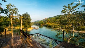 A wooden deck with a hammock and a chair overlooking a river, surrounded by trees and hills.