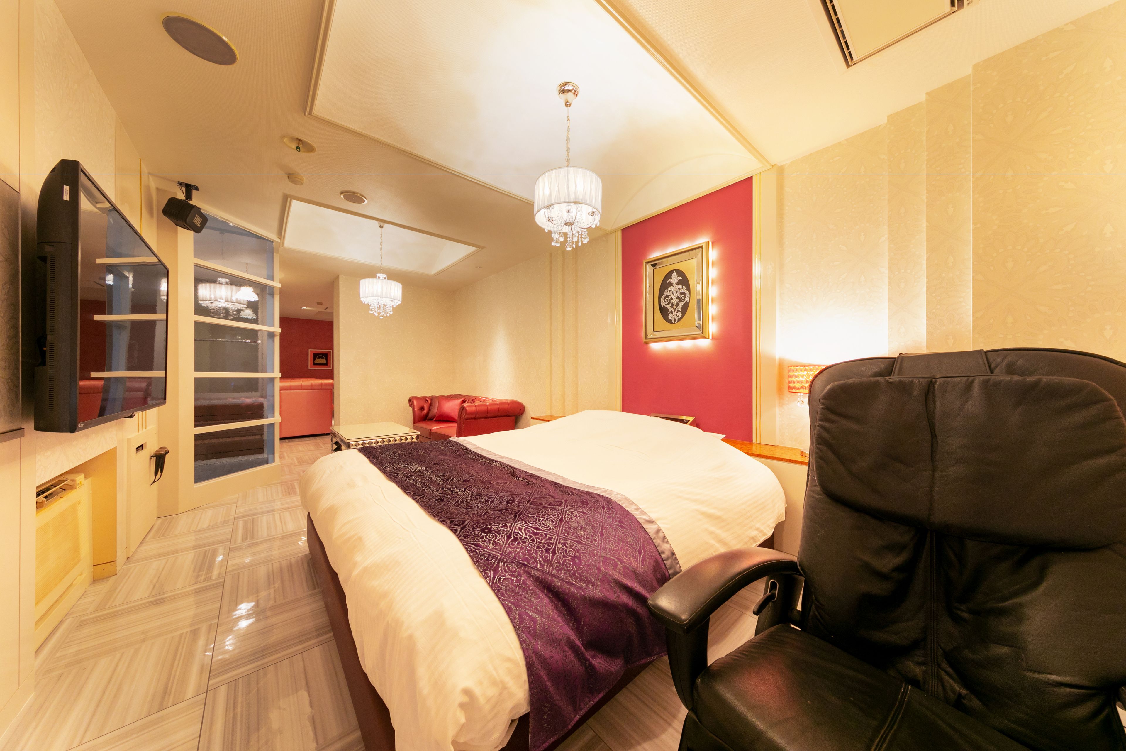 Quarto de hotel com uma cama, uma poltrona de couro preto, uma televisão, um candelabro e uma parede vermelha com um quadro emoldurado.