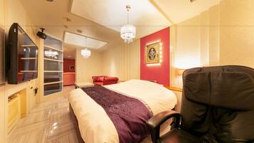 Quarto de hotel com uma cama, uma poltrona de couro preto, uma televisão, um candelabro e uma parede vermelha com um quadro emoldurado.