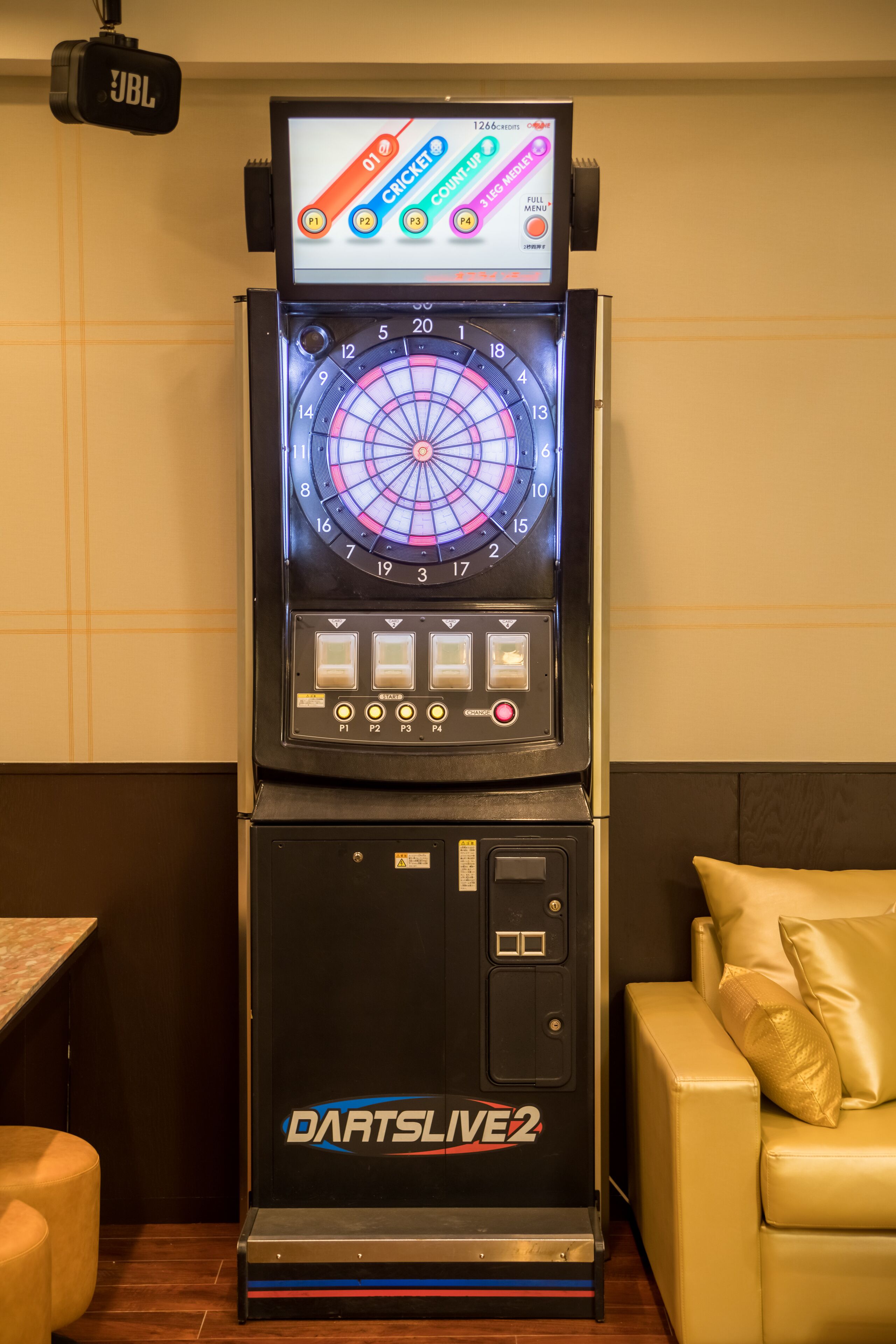 Una máquina de dardos con el letrero "DartsLive 2", que incluye un tablero de dardos, indicador de puntuación y botones de control.